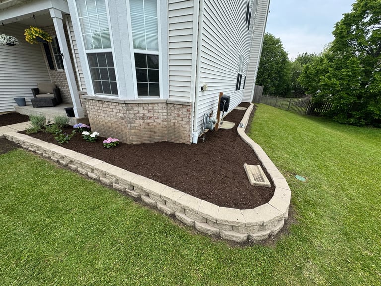 Landscaped garden bed with dark mulch in curved stone border at house foundation against green lawn